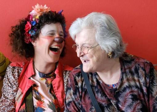 Clown Chouia, fleurs dans les cheveux, fait rire une dame âgée avec des lunettes