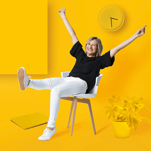 Femme assise sur une chaise qui sourit, pantalon blanc, bras levés, fond jaune