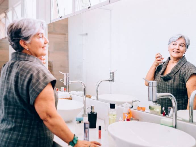 Femme senior, cheveux gris courts, se parfume devant son miroir de salle de bains