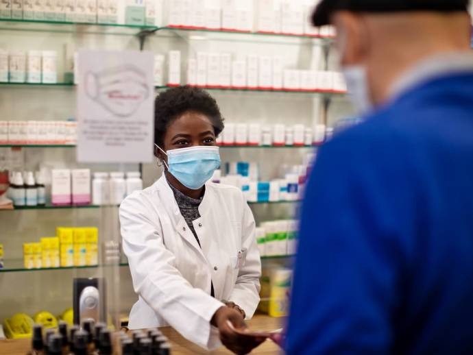 Femme noire masquée, tenue de pharmacienne, prend carte d'un homme de dos avec béret