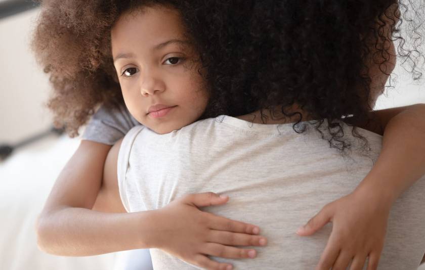 Enfant qui ne sourit pas, serre dans ses bras une adulte vue de dos