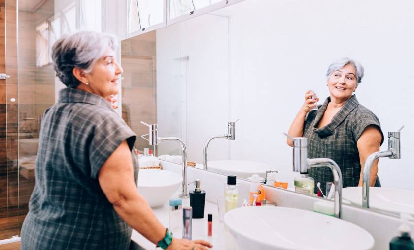 Femme senior, cheveux gris courts, se parfume devant son miroir de salle de bains