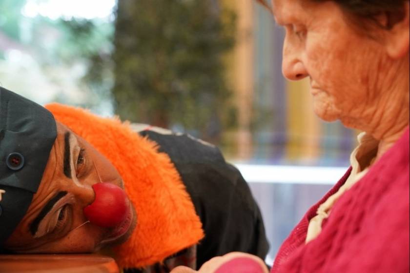 Clown Limengo, col fourrure orange, tête sur une table, regarde une vieille dame triste