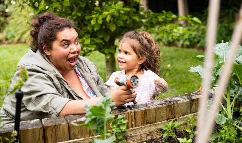 Femme et petite fille rigolent avec tuyau d'arrosage, arrosent des plantes vertes