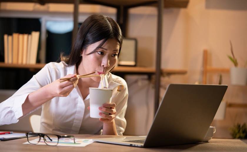 Femme asiatique, mange devant son écran d'ordinateur, papiers sur le bureau