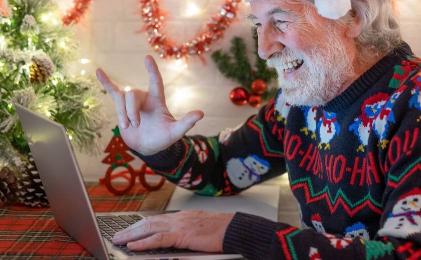 Homme agé, cheveux blancs, avec pull de Noel amusant, sourit devant la caméra de son ordinateur