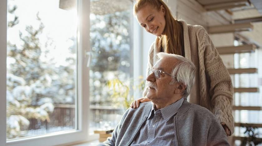 Un homme âgé regarde par la fenêtre, jeune femme souriante lui touche l'épaule