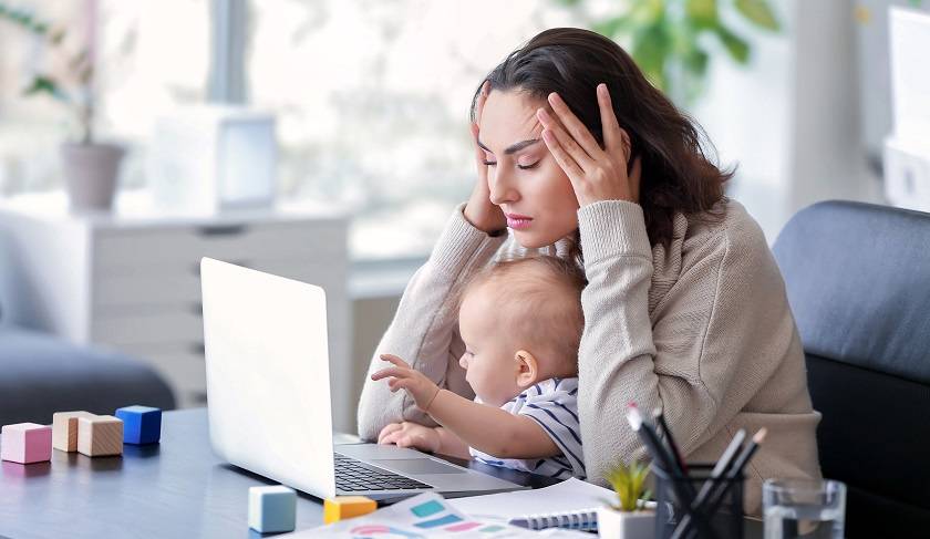 Femme devant un ordinateur et des papiers, tient sa tête entre ses mains, bébé sur ses genoux