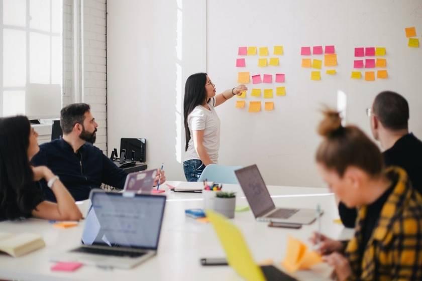 Réunion d'équipe au travail, avec plusieurs personnes autour d'une table et une femme au tableau