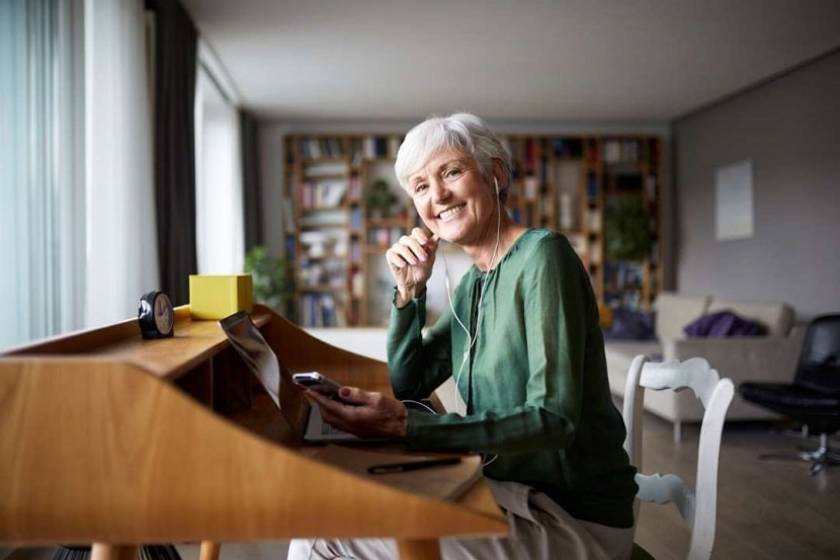 Une senior est assise devant son bureau, écouteurs aux oreilles et téléphone portable à la main